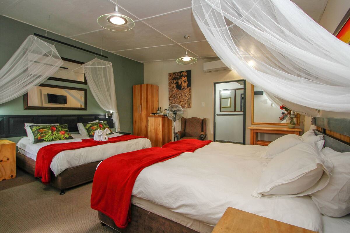A hotel room with two beds, each with white bedding and red throw blankets, a wooden wardrobe, a fan, and a large mirror.