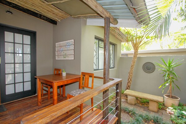 A wooden dining table with chairs, a bench, and a potted plant in a shaded outdoor area.