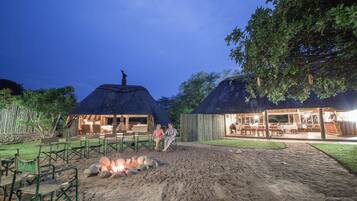 A thatched-roof outdoor dining area with a fire pit, wooden furniture, and a view of the surrounding landscape.