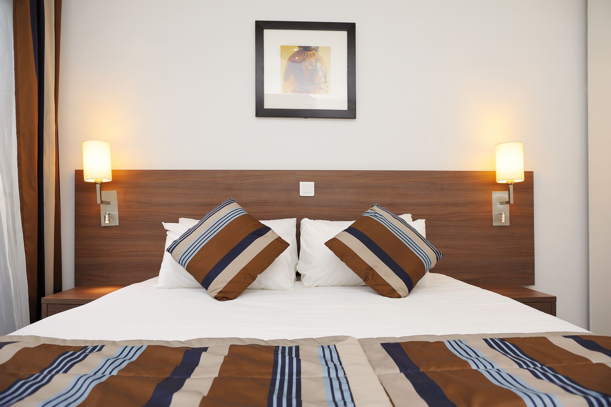 A hotel room with a large bed, two bedside lamps, a framed picture on the wall, and a window with curtains.