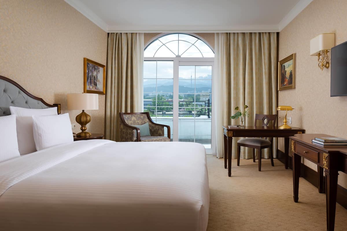 A hotel room with a large bed, a desk, a chair, a lamp, a television, and a view of the city through a large window.