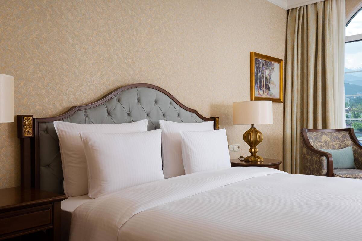 A hotel room with a large bed, a nightstand, a lamp, a chair, and a painting on the wall.