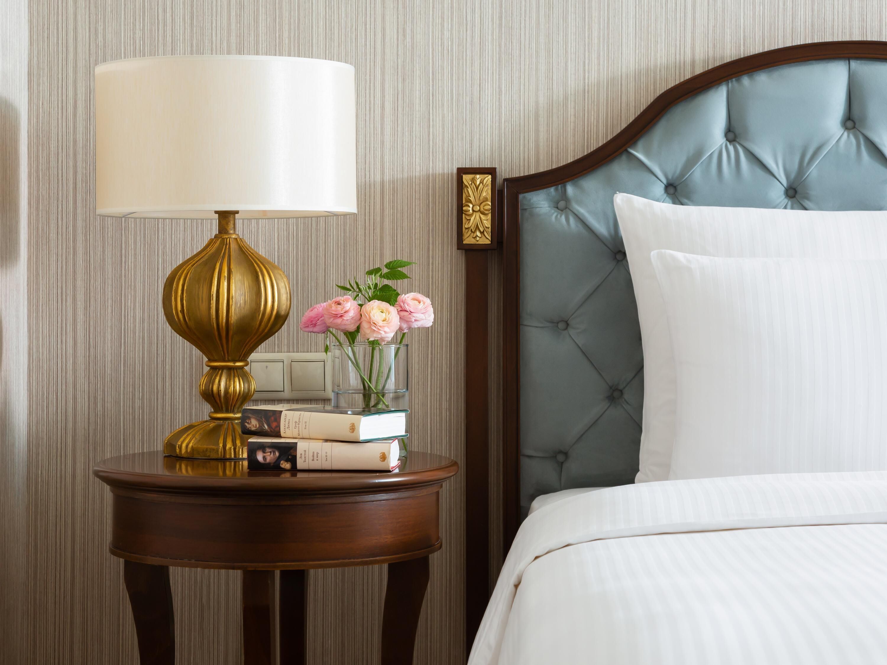 A neatly made bed with a tufted headboard, a bedside table with a lamp, books, and a vase of flowers.