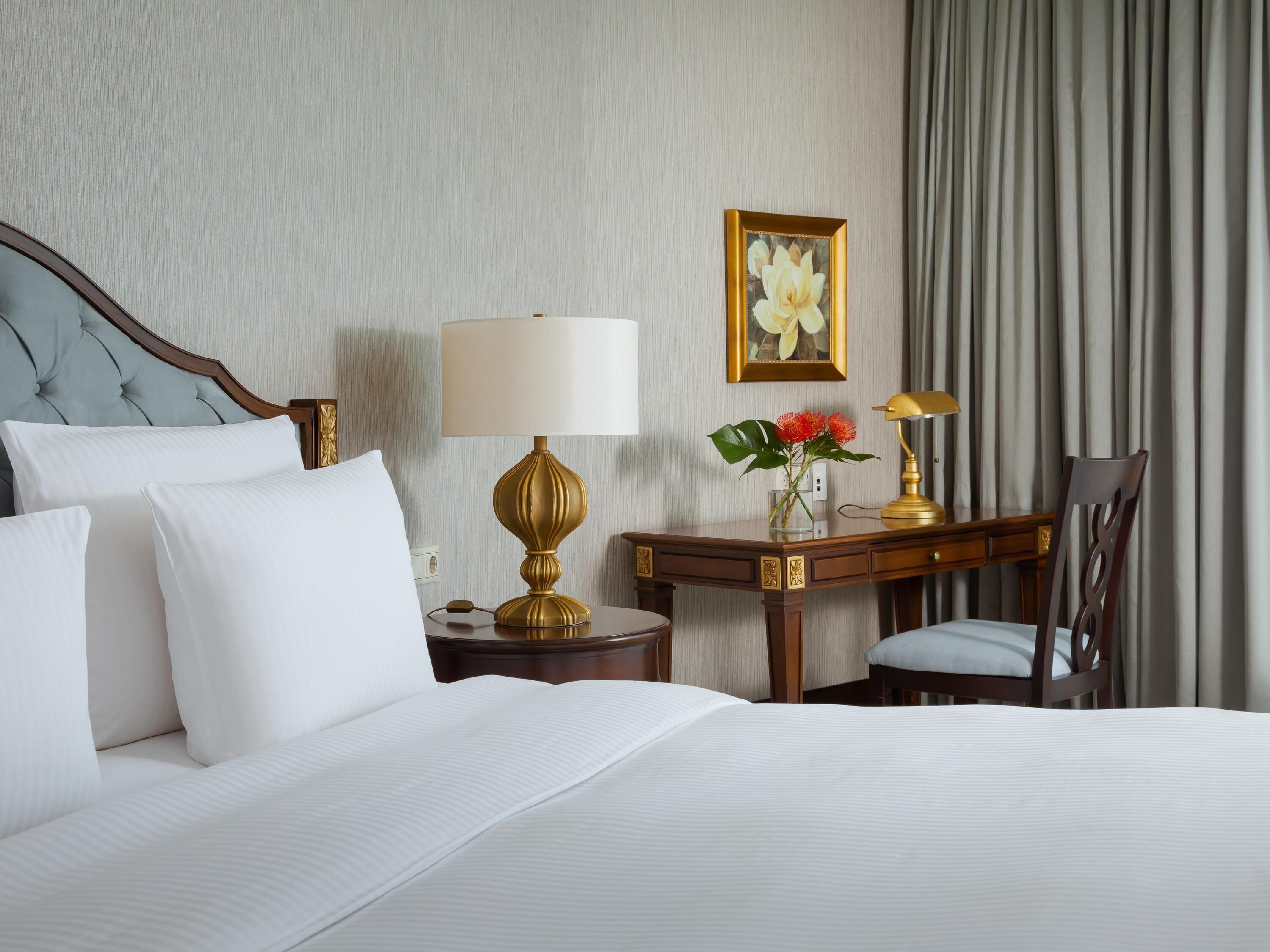 A hotel room with a bed, a desk with a lamp, a chair, and a picture on the wall.