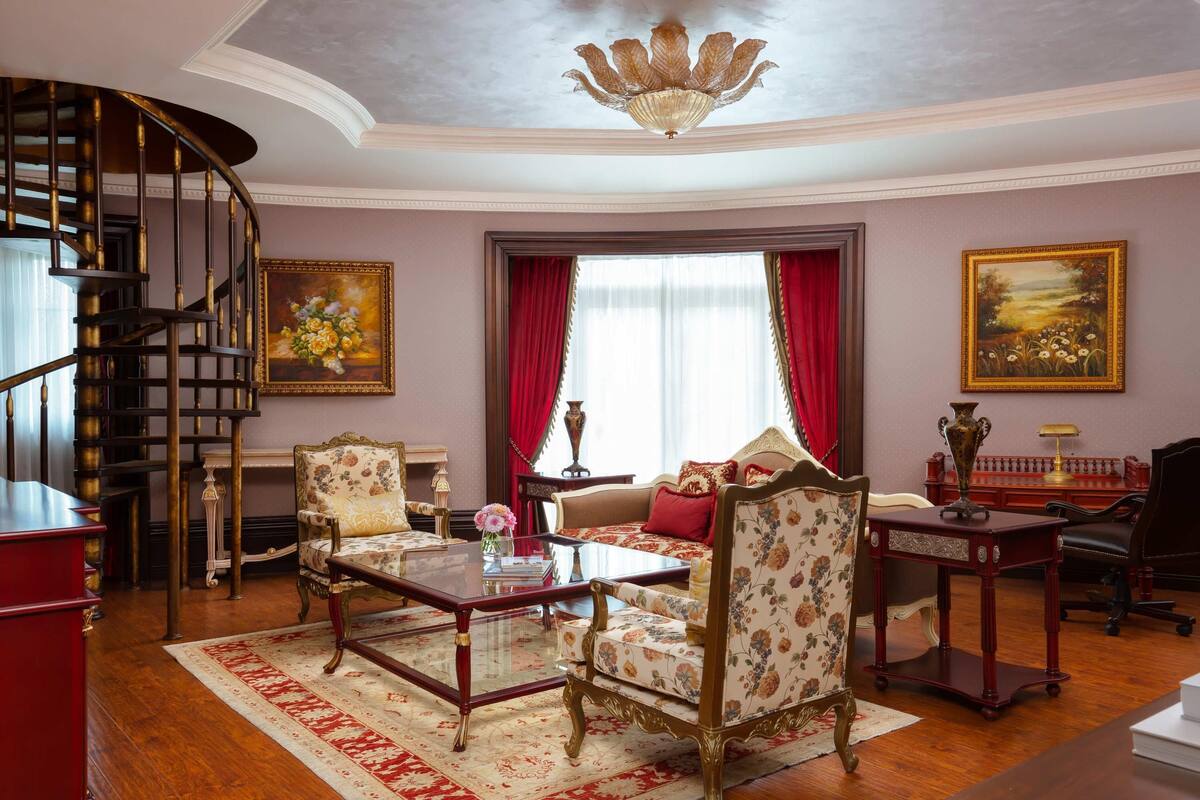A room with a wooden floor, a red and white patterned rug, a round table, two chairs, a sofa, a desk, a chair, a bookshelf, a painting, a chandelier, and a spiral staircase.