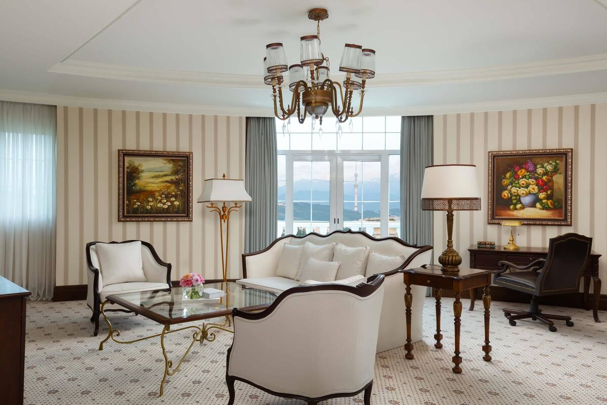 Quarto de hotel com uma grande janela, um candelabro, um sofá, uma mesa de escritório e duas cadeiras.