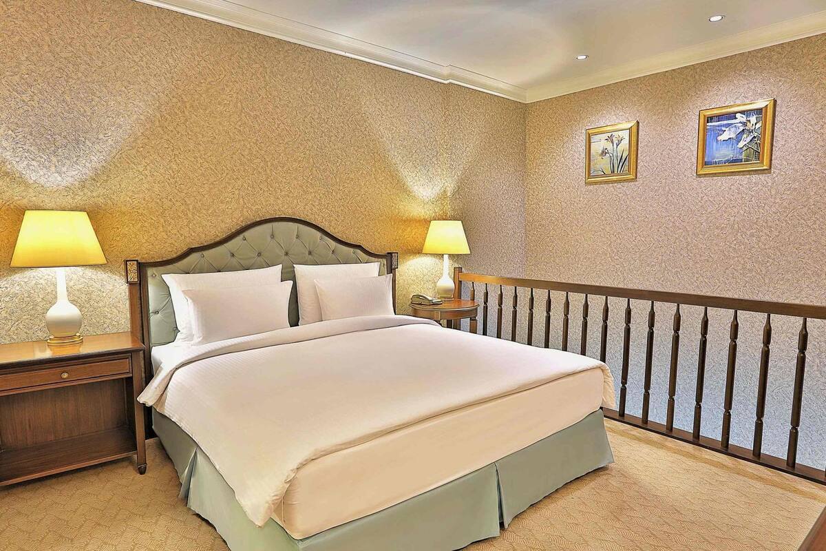 A hotel room with a large bed, two bedside tables with lamps, a wooden headboard, and a patterned carpet.