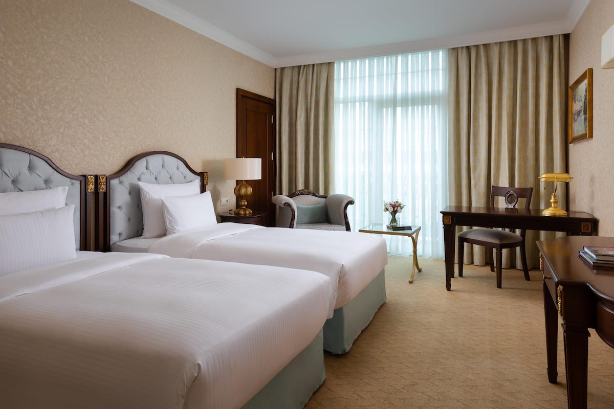 A hotel room with two beds, a desk, a chair, a small table, and a large window with curtains.