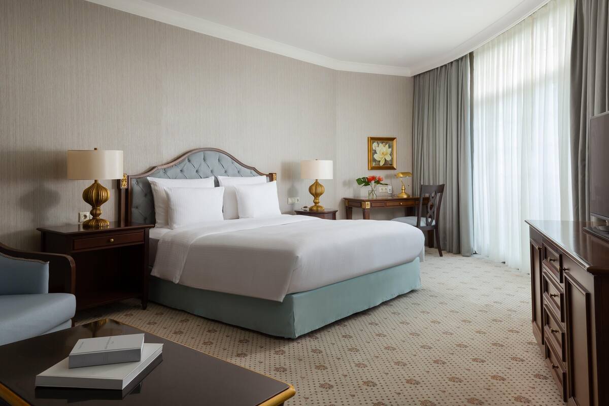 A hotel room with a bed, two bedside tables with lamps, a desk with a chair, a television, and a large window with curtains.