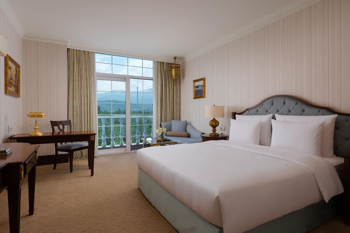 A hotel room with a large bed, a desk, a chair, a nightstand, and a view of mountains through a large window.