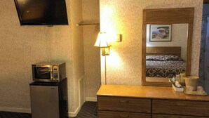 A hotel room with a flat-screen TV, a microwave, a refrigerator, a wooden dresser, a bedside lamp, and a framed picture on the wall.