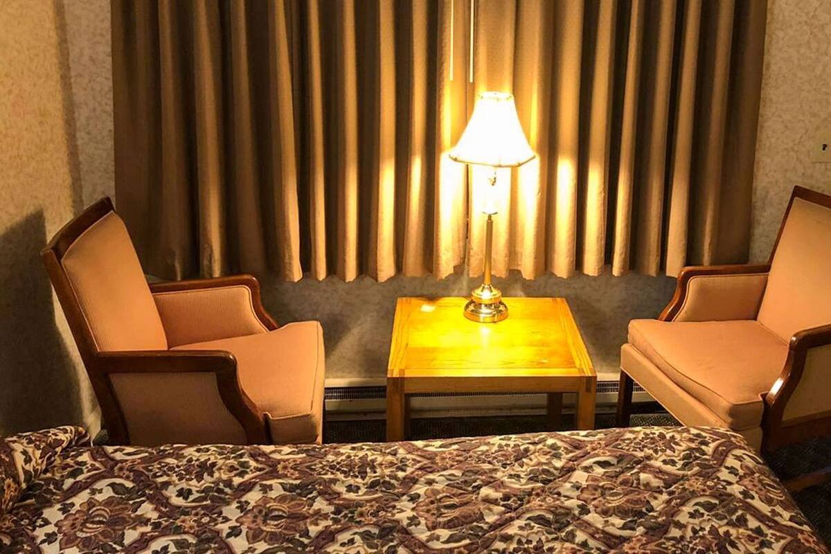 Ein Hotelzimmer mit einem Bett, zwei Sesseln, einem kleinen Tisch, einer Lampe und einem Fenster mit vertikalen Jalousien.