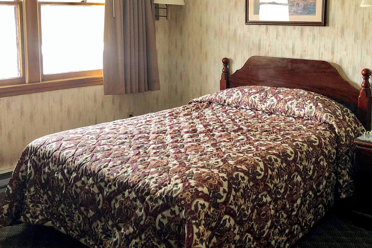 Ein Hotelzimmer mit einem Bett, einem Fenster, einer Lampe und einem gerahmten Bild an der Wand.