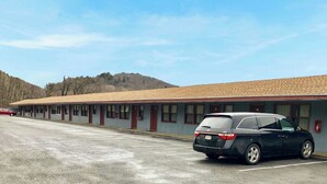Un motel con una fila de habitaciones, un estacionamiento y un paisaje montañoso al fondo.