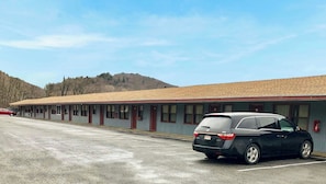 Un motel con una fila de habitaciones, un estacionamiento y un paisaje montañoso al fondo.