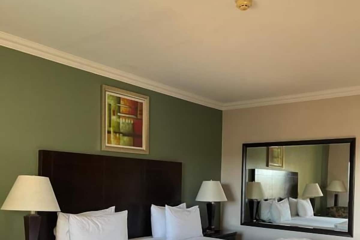 Una habitación de hotel con una cama grande, dos mesitas de noche con lámparas, un cabecero, un espejo grande y un cuadro en la pared.