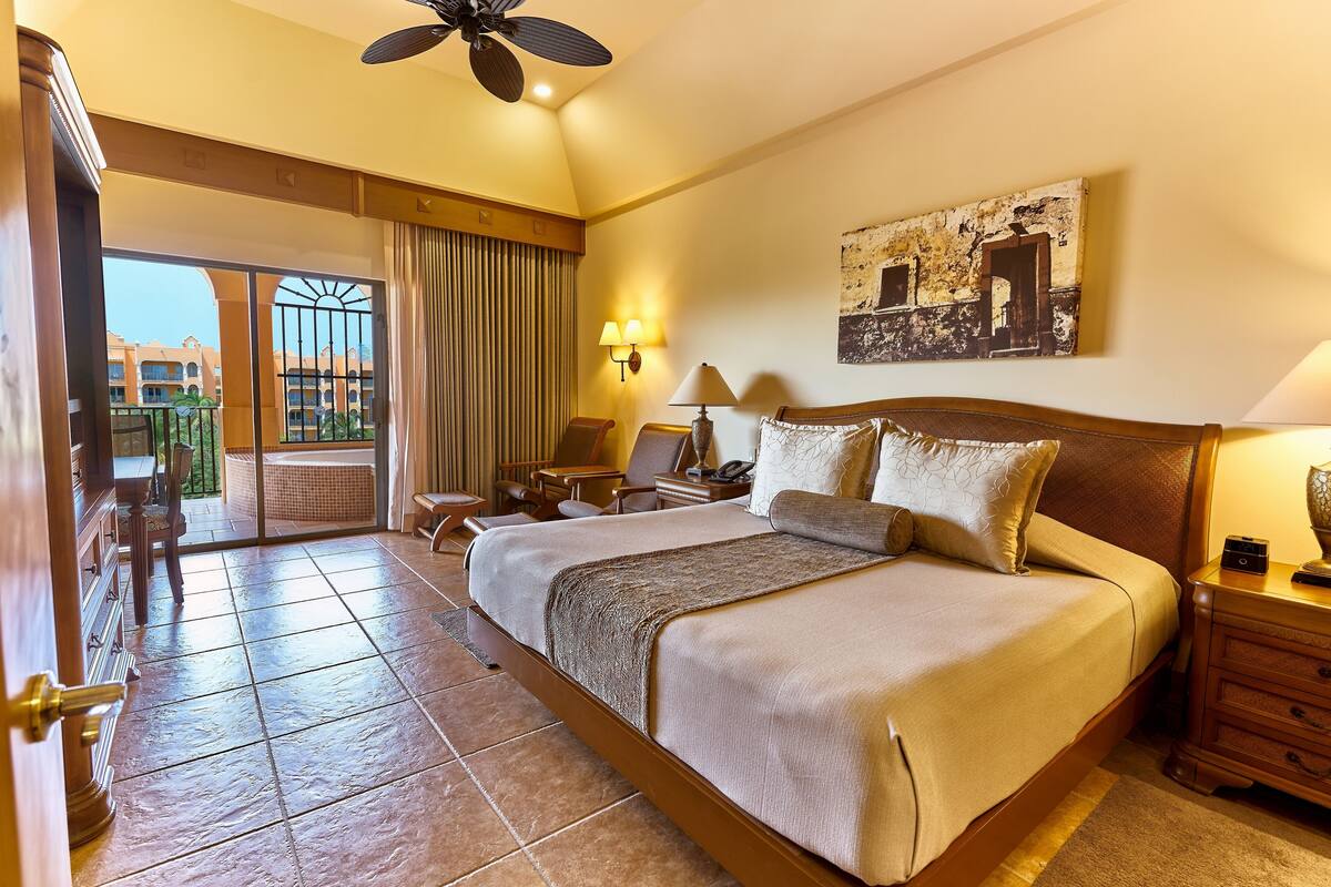 A hotel room with a large bed, a ceiling fan, a balcony with a view of other buildings, and a painting on the wall.