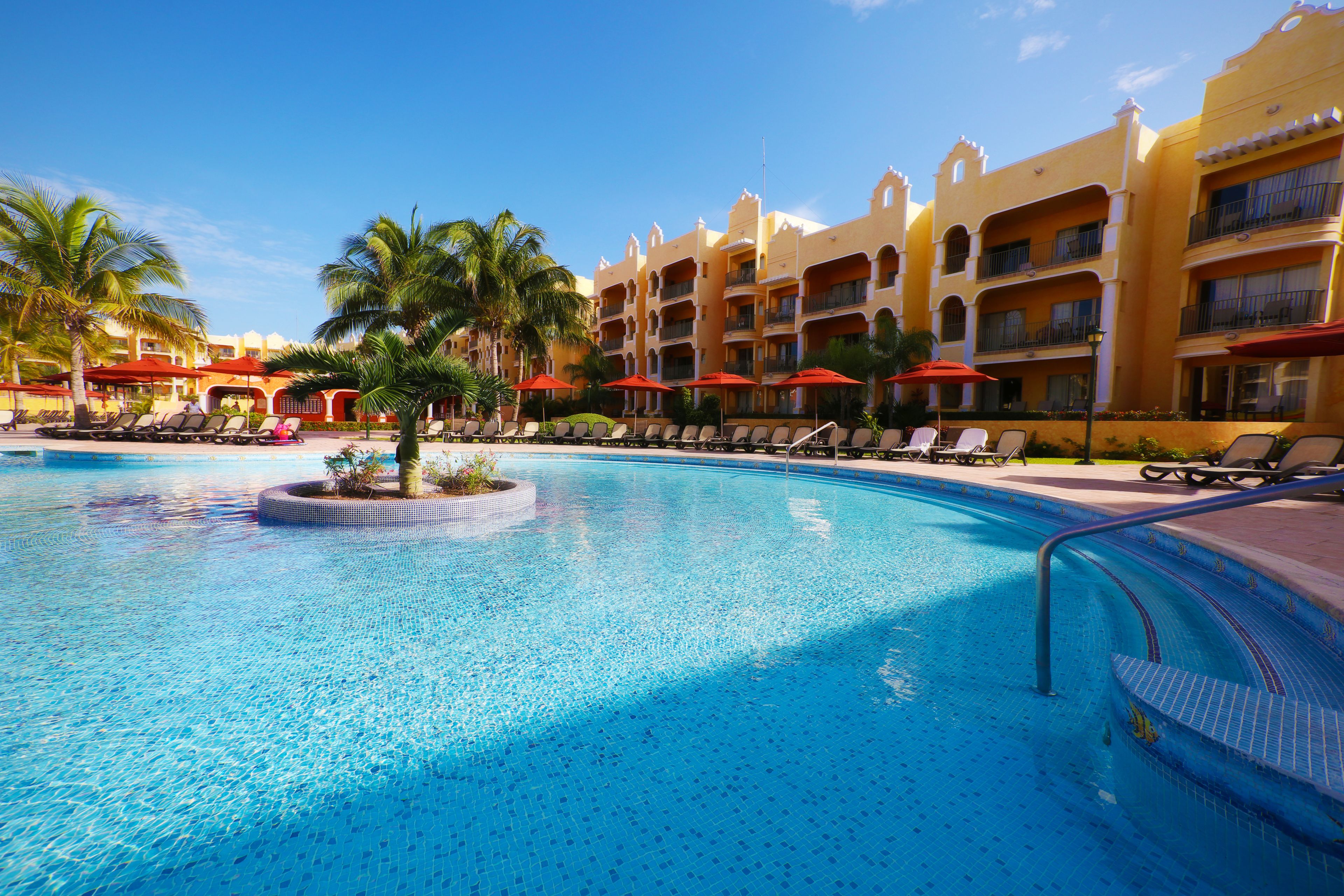 Piscina con sillones de descanso y sombrillas, rodeada de palmeras y un edificio de hotel con balcones.