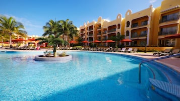Piscina con sillones de descanso y sombrillas, rodeada de palmeras y un edificio de hotel con balcones.