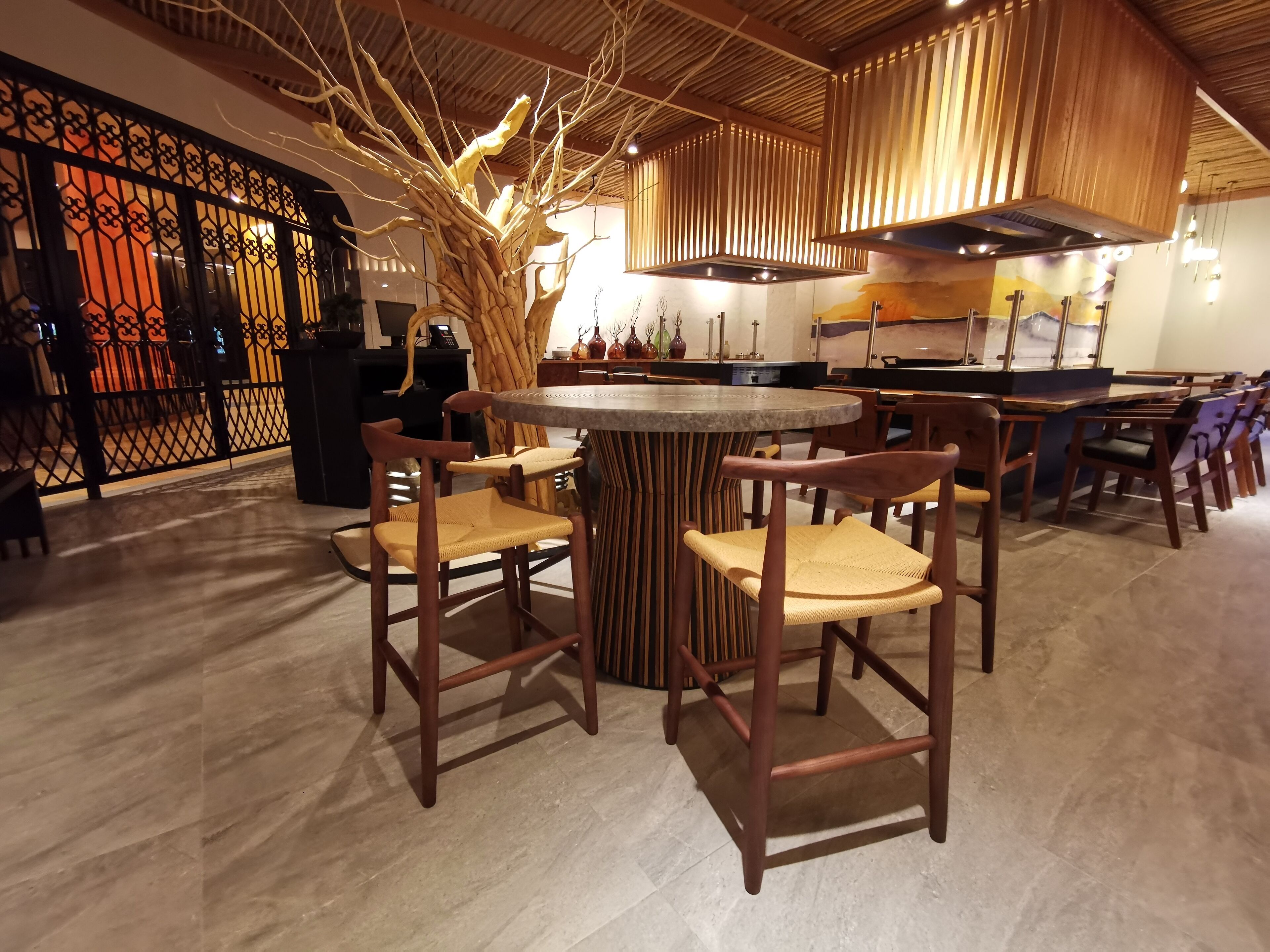Interior de restaurante com uma mesa redonda central rodeada por cadeiras de madeira, uma escultura de árvore decorativa e uma cozinha moderna ao fundo.