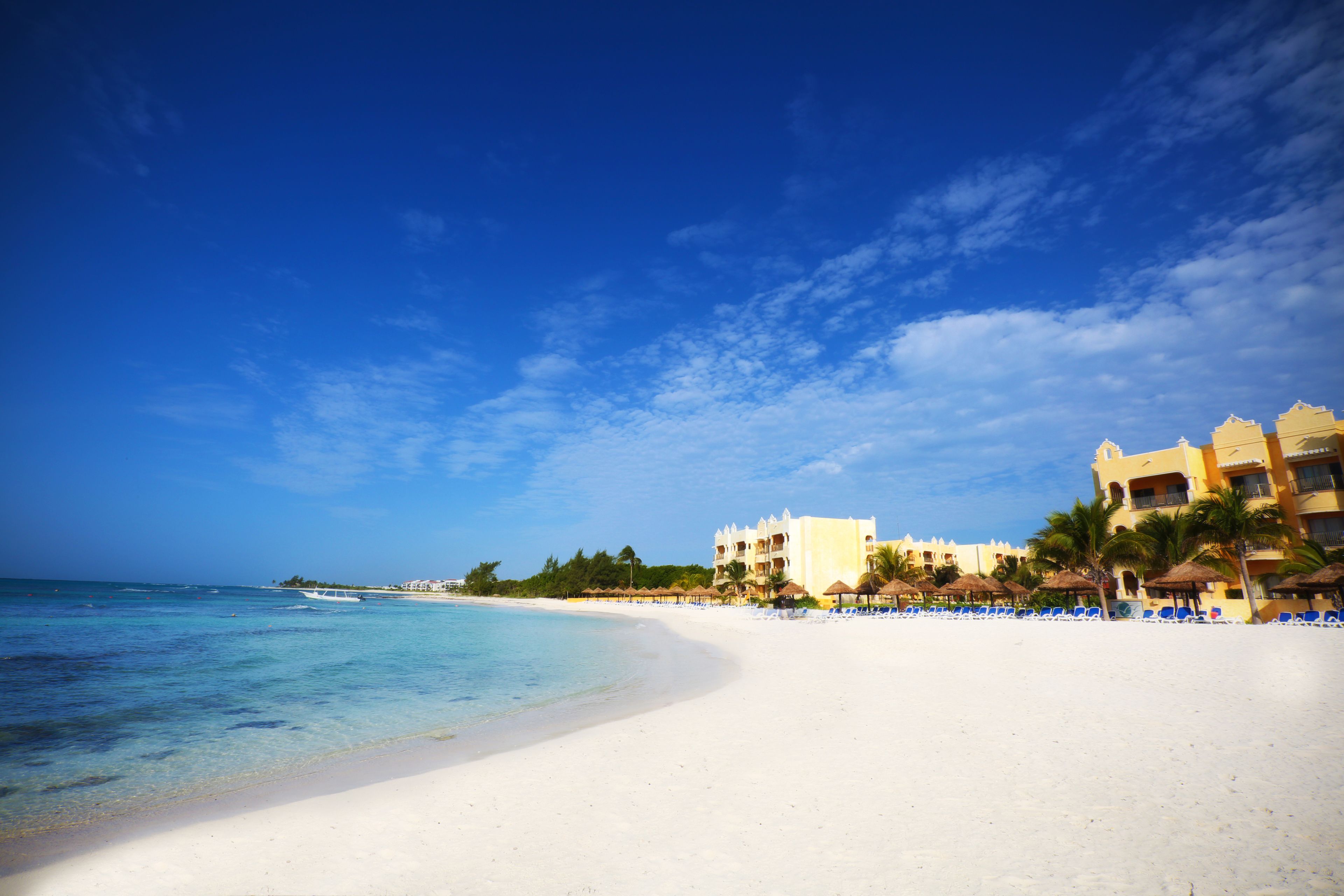 Ein Strand mit weißem Sand, klarem, blauem Wasser und einem Resorthaus mit mehreren Balkonen und Palmen.