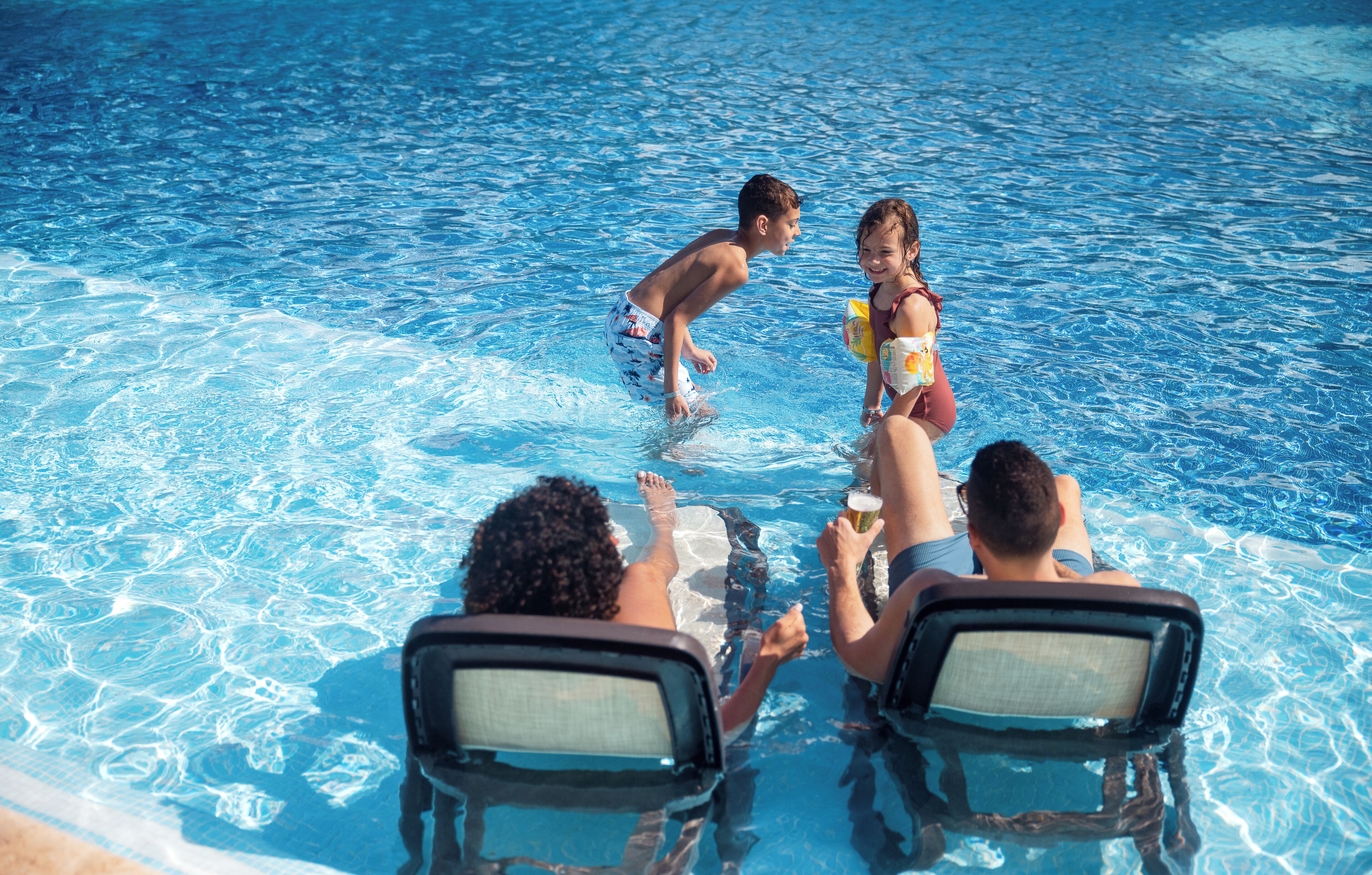 A swimming pool with clear blue water, a few people in the water, and two empty pool chairs.