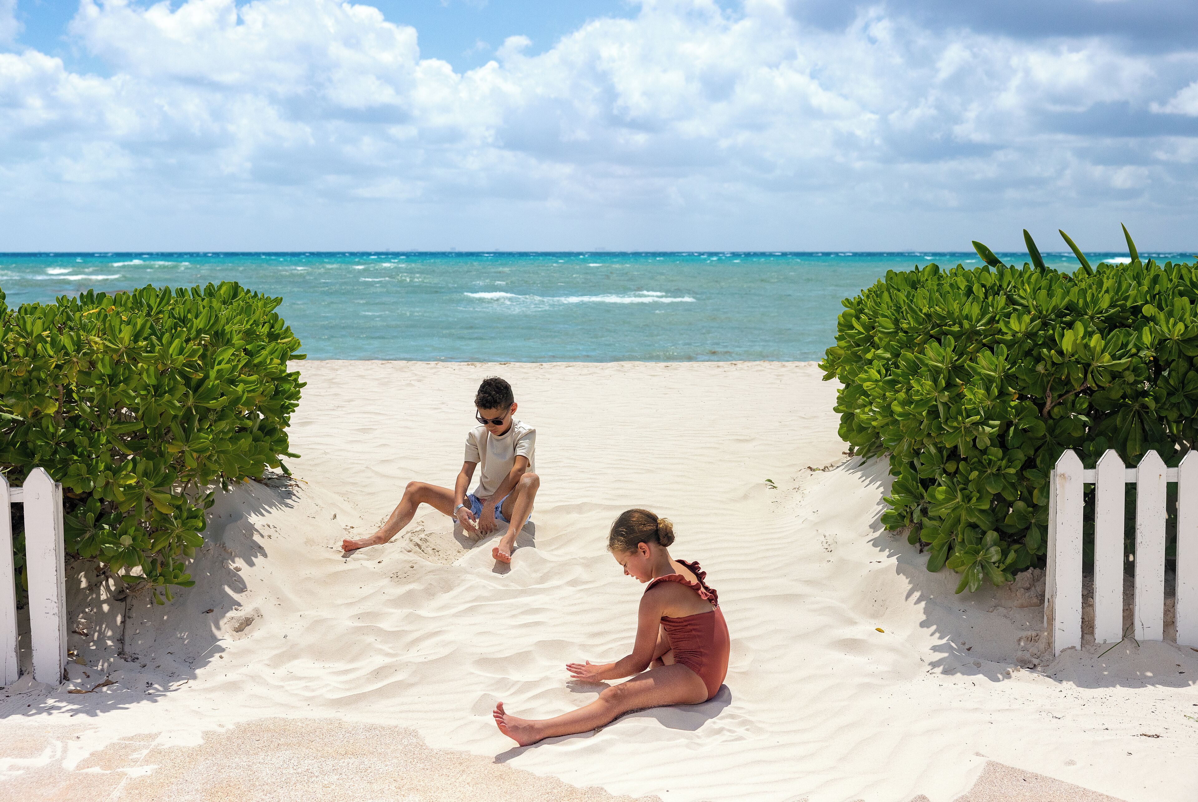 A sandy beach with a white picket fence and green bushes, leading to the ocean.
