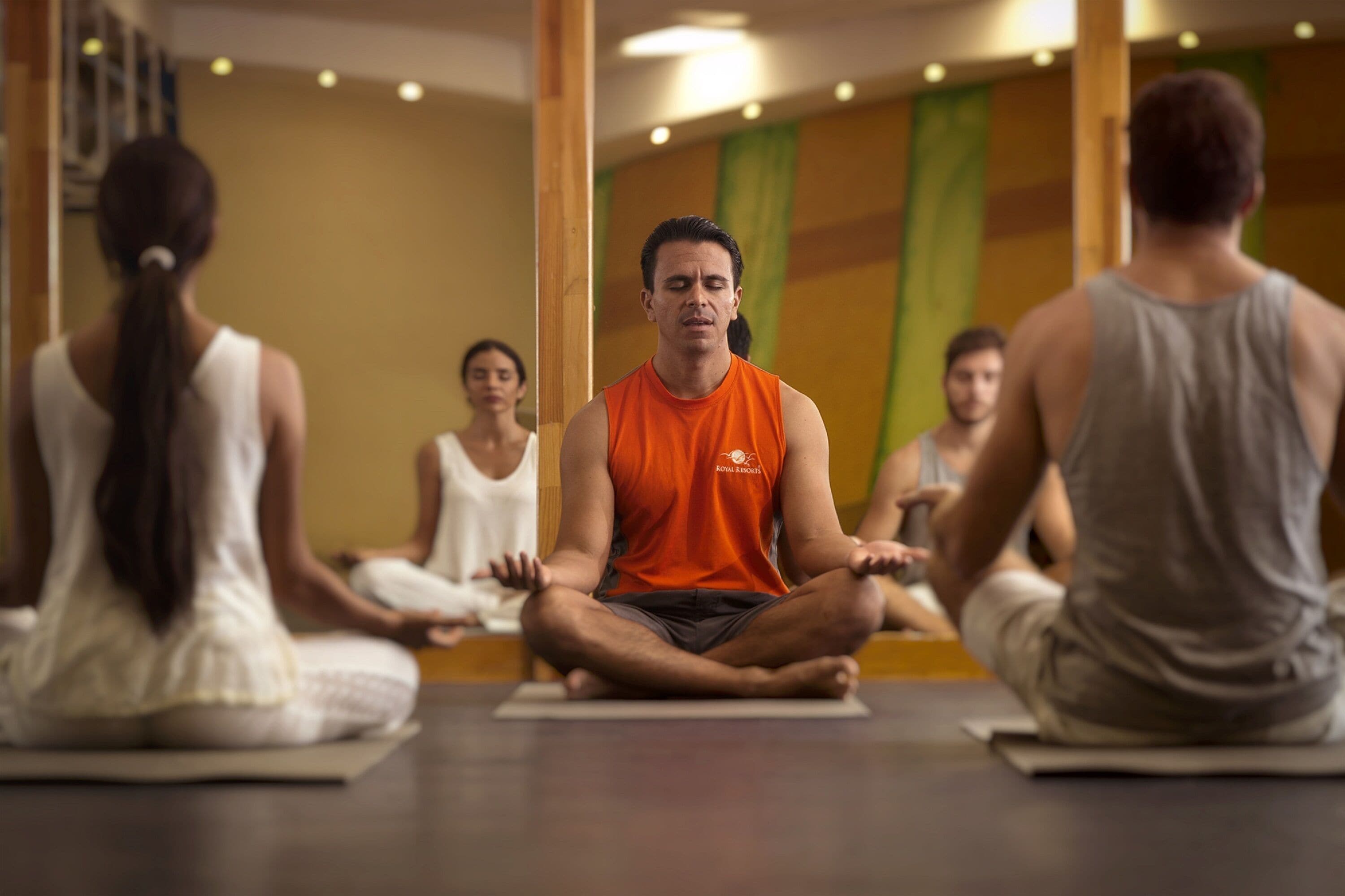 A group of people sitting cross-legged on yoga mats in a room with wooden pillars and a mirror on the wall.