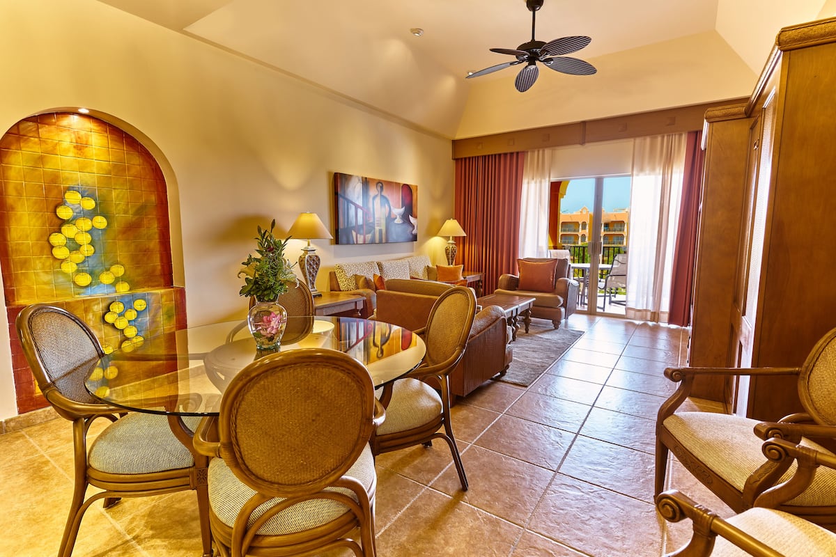 A hotel room with a dining area, a large window with curtains, a ceiling fan, and a decorative wall piece.