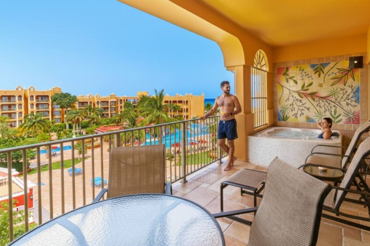A balcony with a view of a pool and resort buildings, furnished with chairs and a table.
