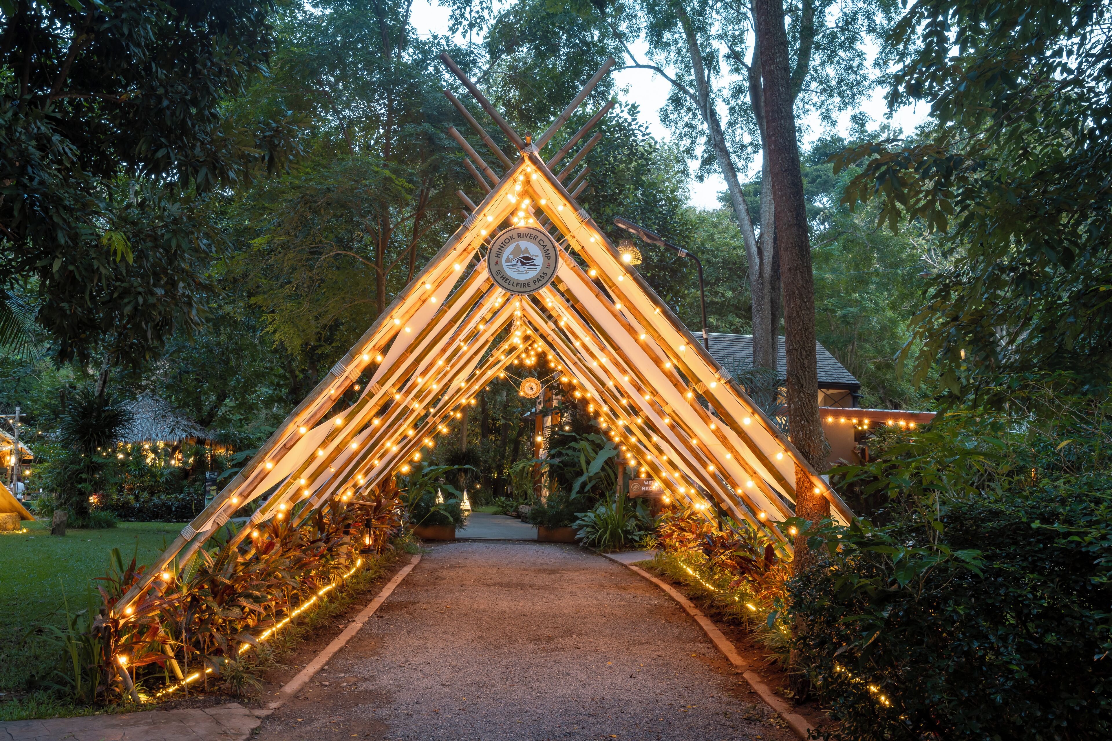 Um caminho serpenteia por baixo de um arco triangular decorado com luzes de corda, rodeado por vegetação exuberante.