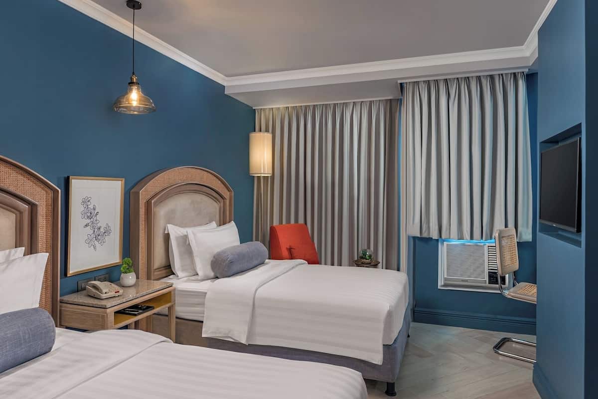 A hotel room with two beds, a desk, a lamp, a TV, and a window with curtains.