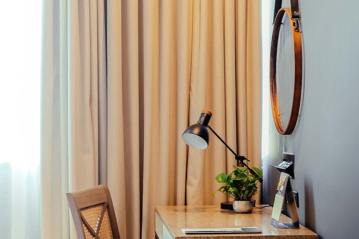 A hotel room with a wooden desk, a chair, a lamp, a potted plant, a mirror, and a bed.