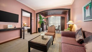 A hotel room with a flat-screen TV, a coffee table with a flower vase, a sofa, a chair, a dining table with chairs, a potted plant, and a large window with curtains. - Makati Palace Hotel (Makati)