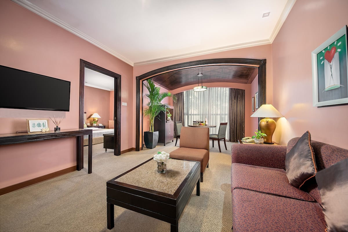 A hotel room with a flat-screen TV, a coffee table with a flower vase, a sofa, a chair, a dining table with chairs, a potted plant, and a large window with curtains.
