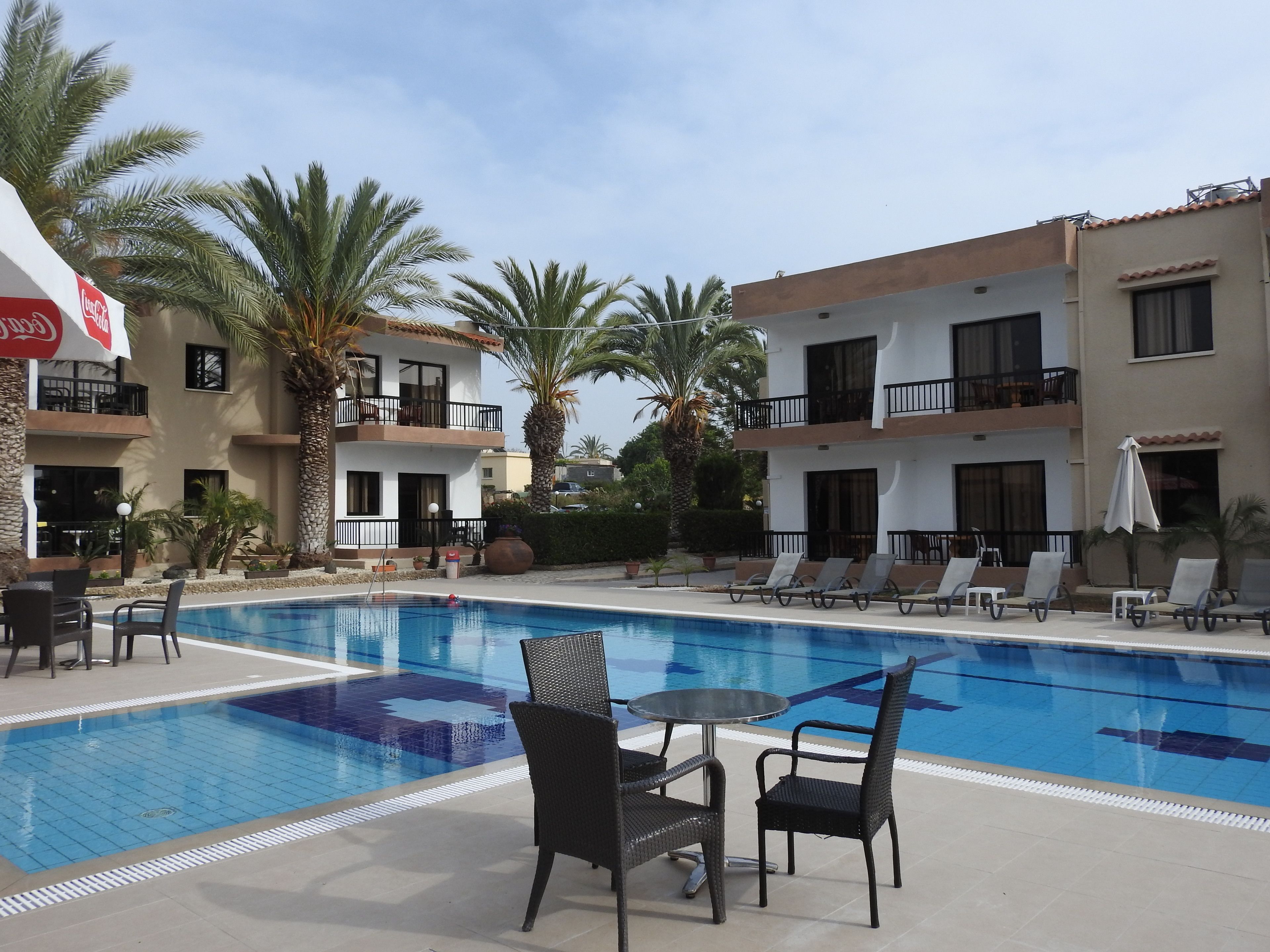Zona de piscina amb gandules i taules, envoltada de palmeres i un edifici amb balcons.