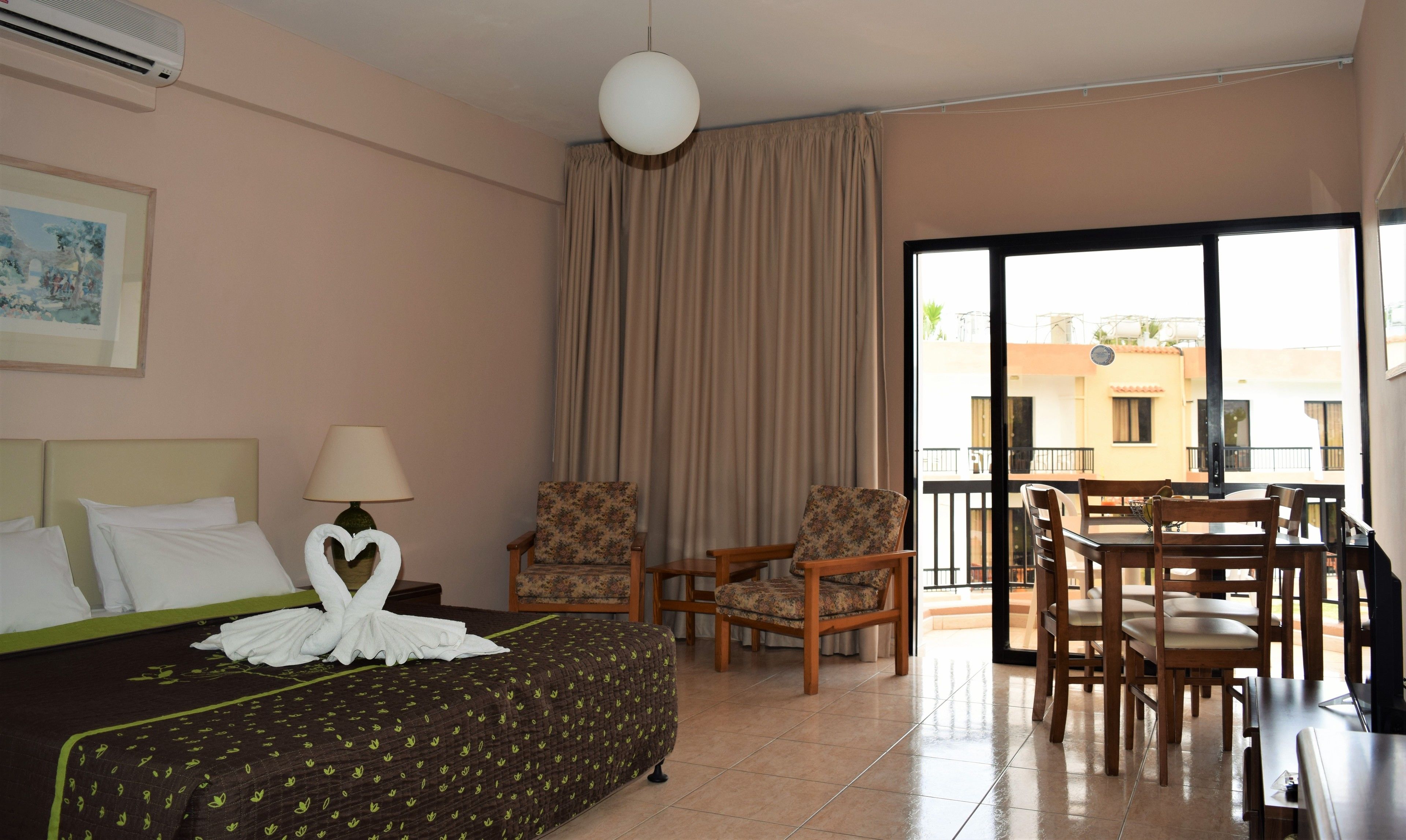 A hotel room with a bed, two chairs, a table, a lamp, and a large window with a view of buildings.