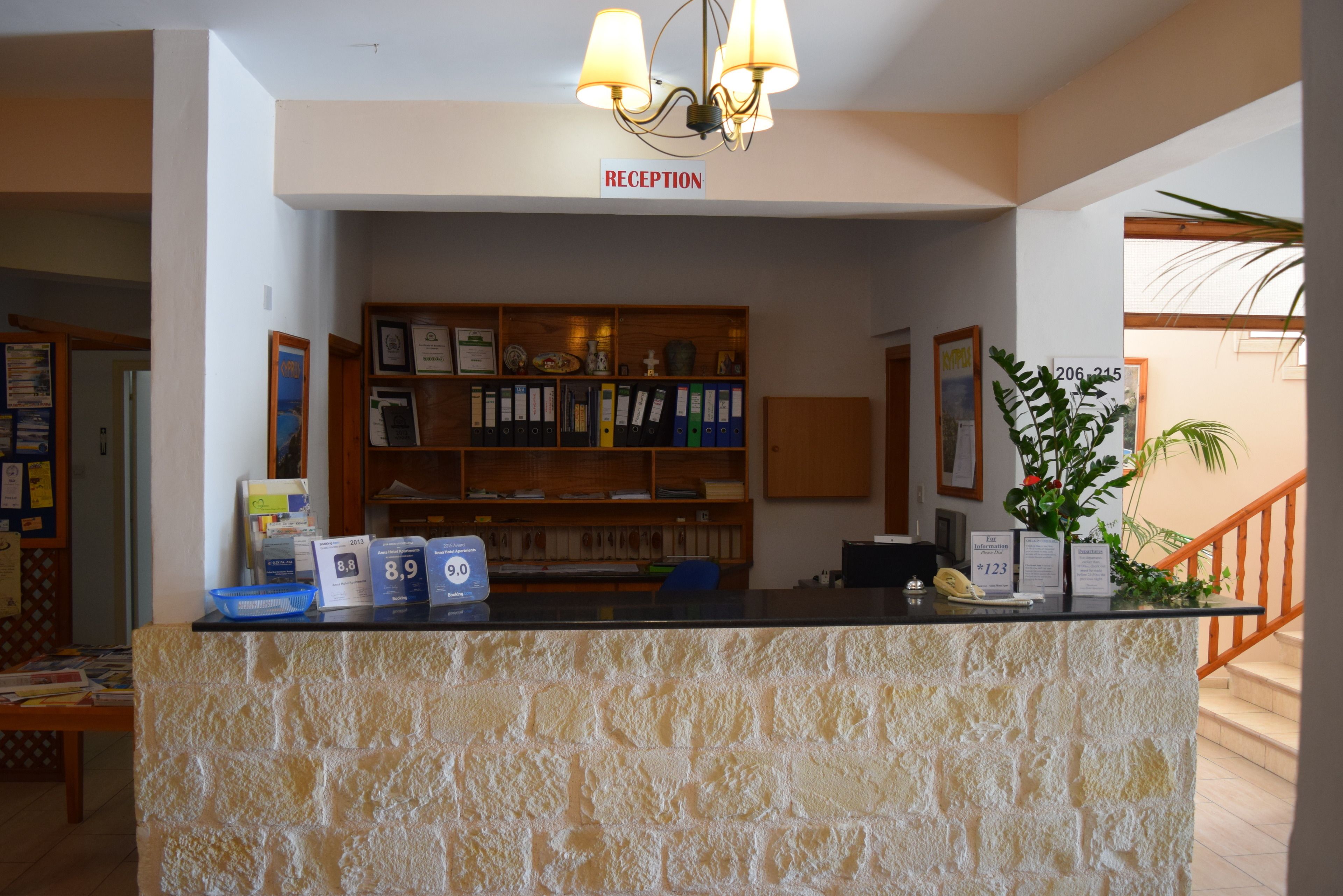 Un área de recepción con mostrador de piedra, una araña, un letrero de recepción, una planta y una escalera.
