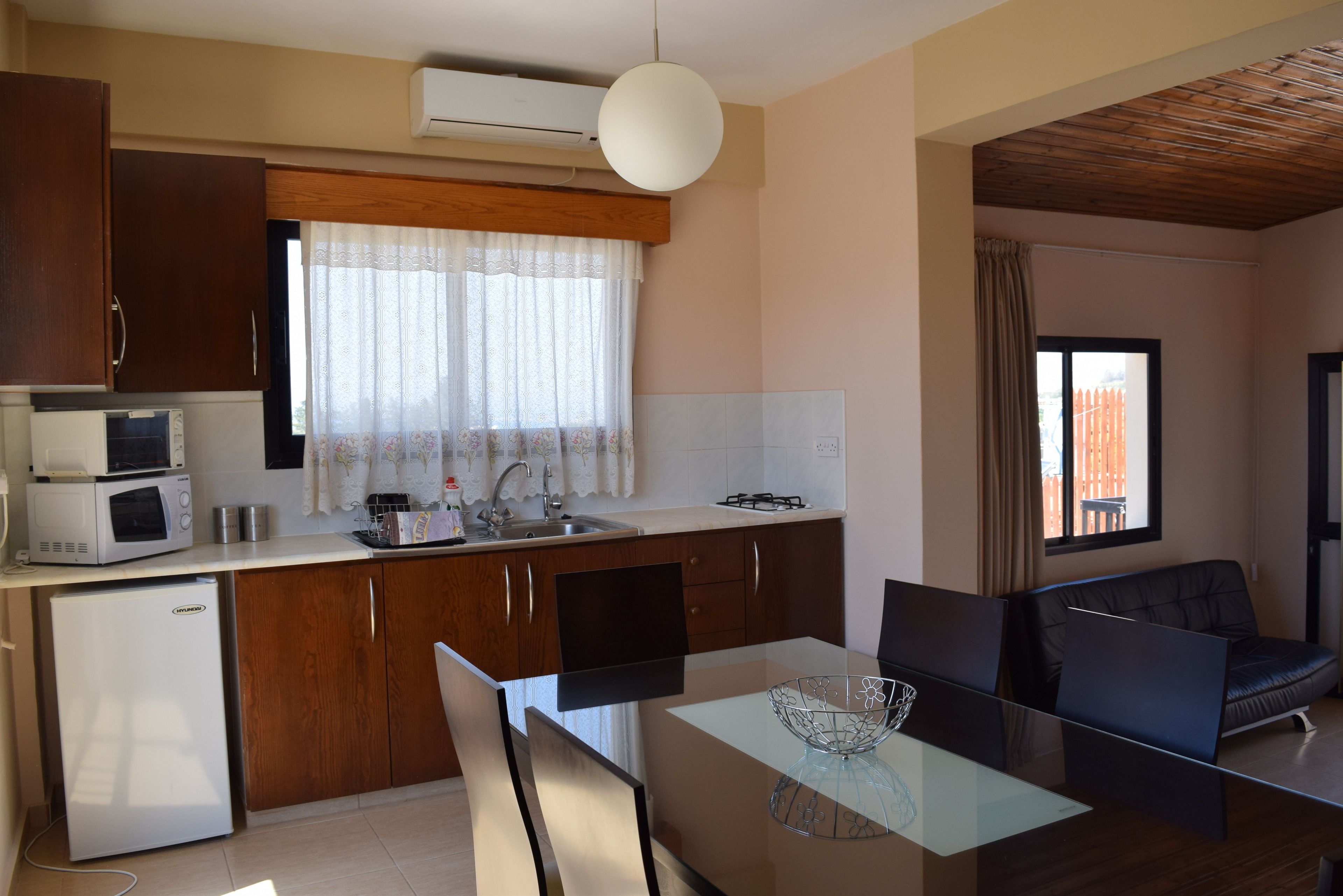 A compact kitchen with a refrigerator, microwave, and stove. There is a dining area with a glass table and chairs, and a sofa.