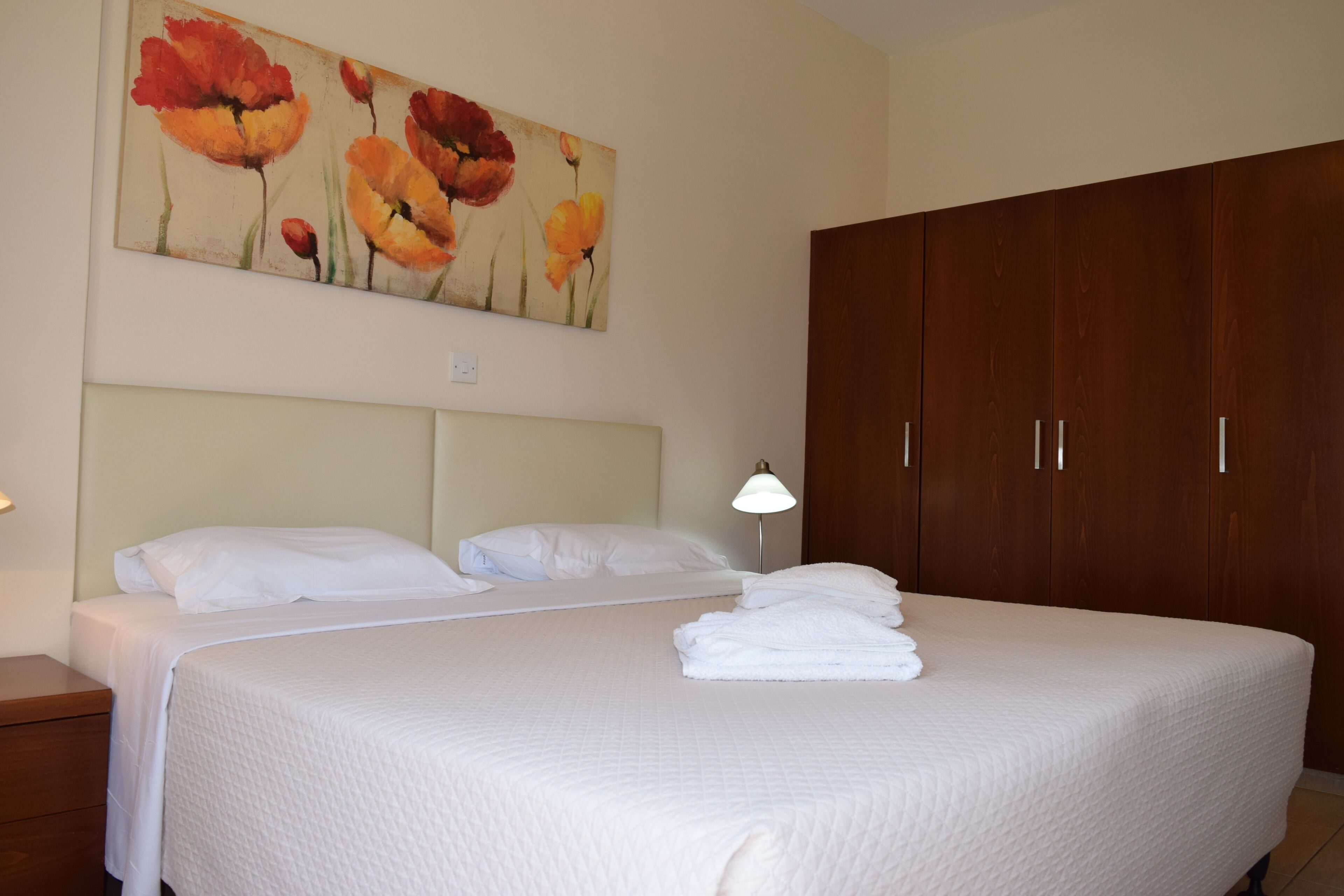 A hotel room with a large bed, a wooden wardrobe, a painting of flowers on the wall, and a bedside lamp.