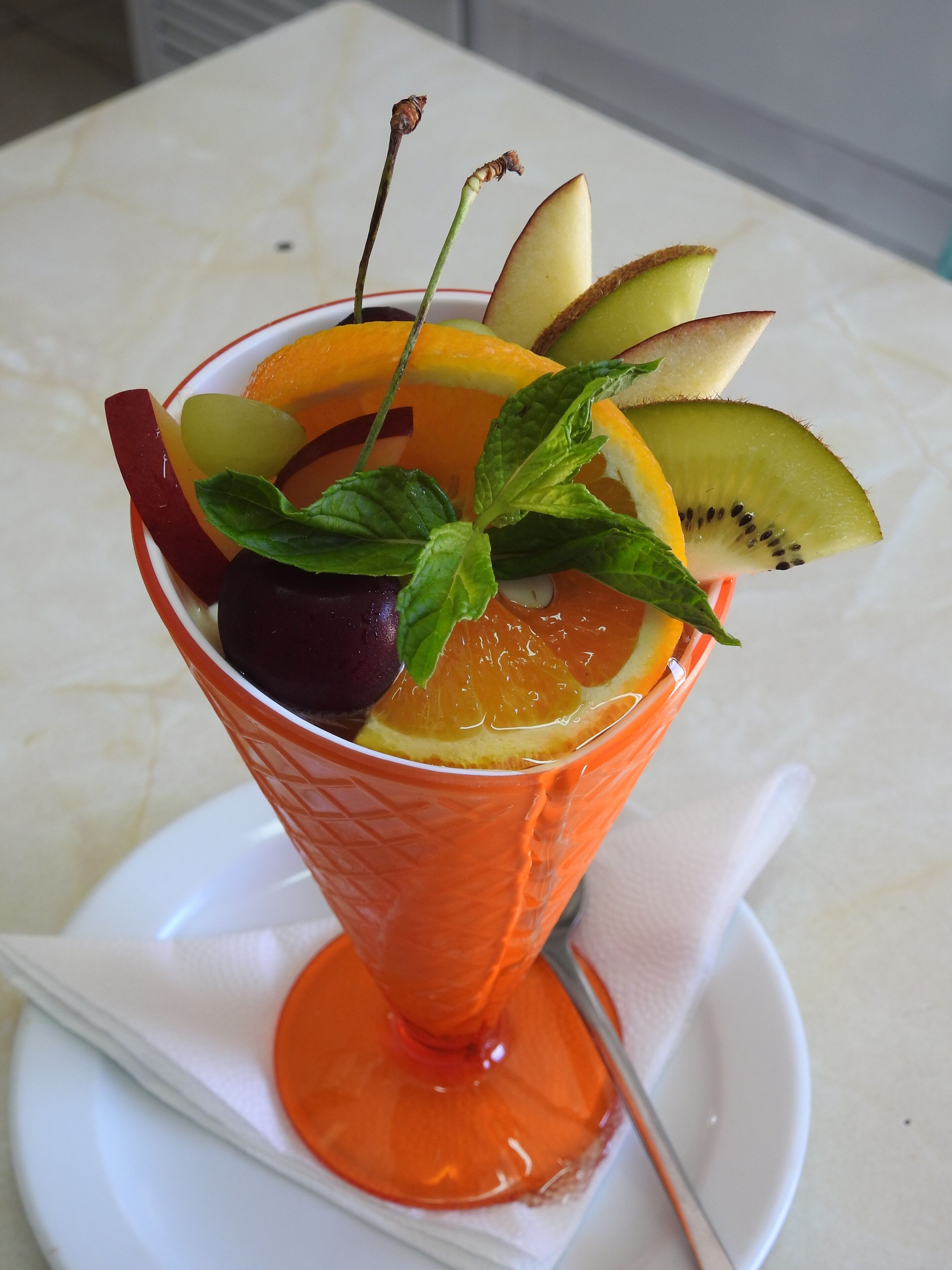 Ein fruchtiger Cocktail in einem verzierten Glas auf einem weißen Teller mit einem Löffel.