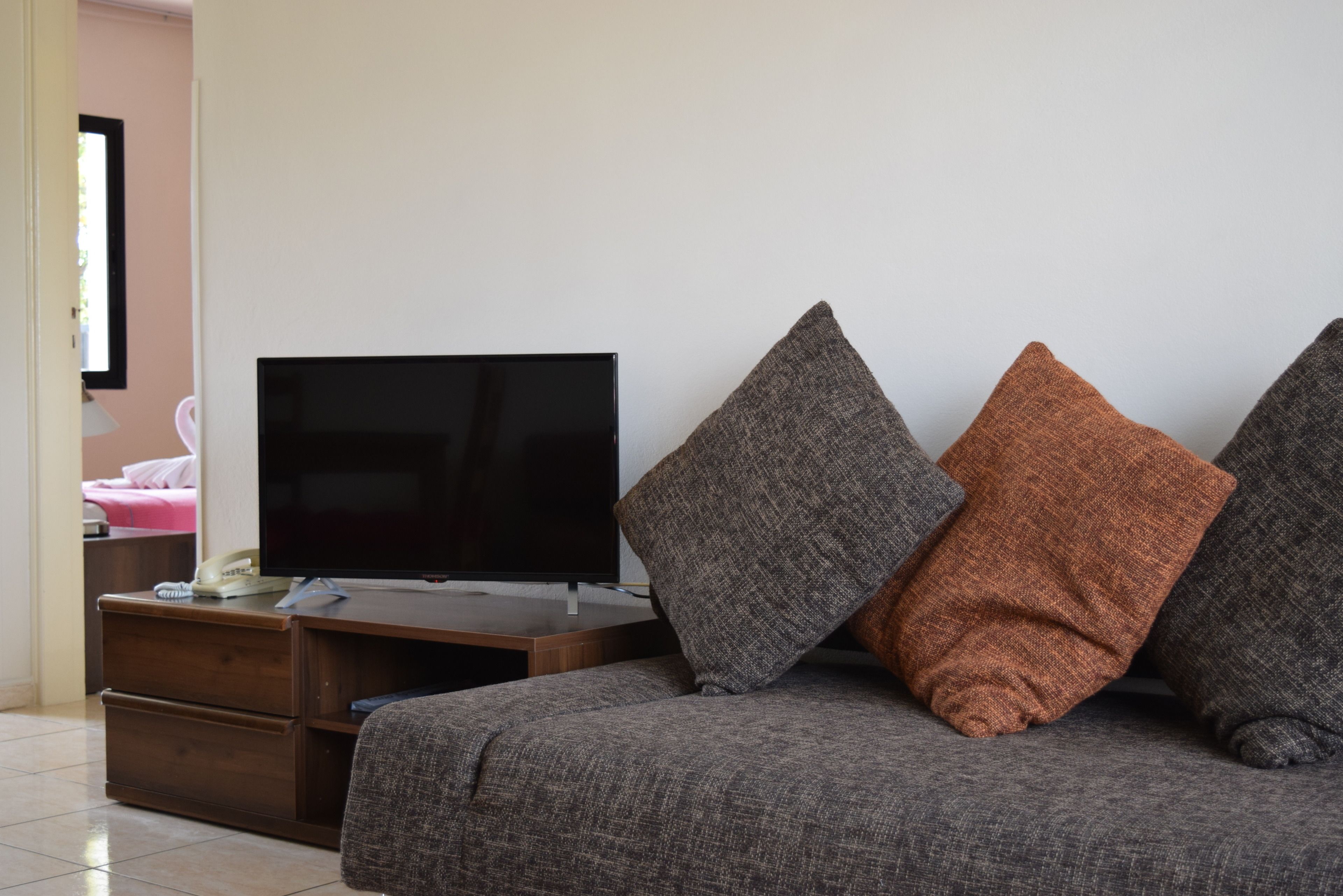 Una sala de estar con una televisión de pantalla plana sobre un soporte de madera, un sofá gris con tres cojines y una silla rosa al fondo.