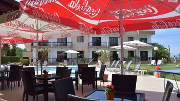 An outdoor seating area with tables and chairs, red umbrellas, and a pool in the background.