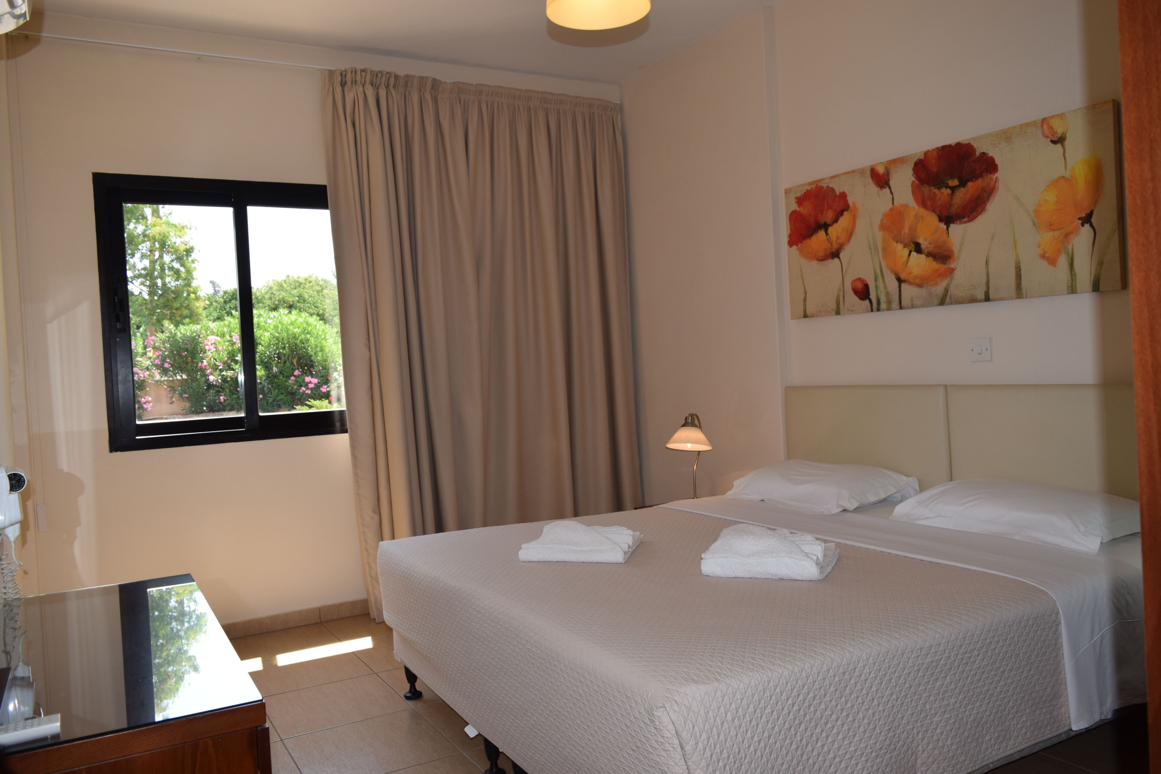 A hotel room with a bed, a window with curtains, a lamp, and a painting on the wall.