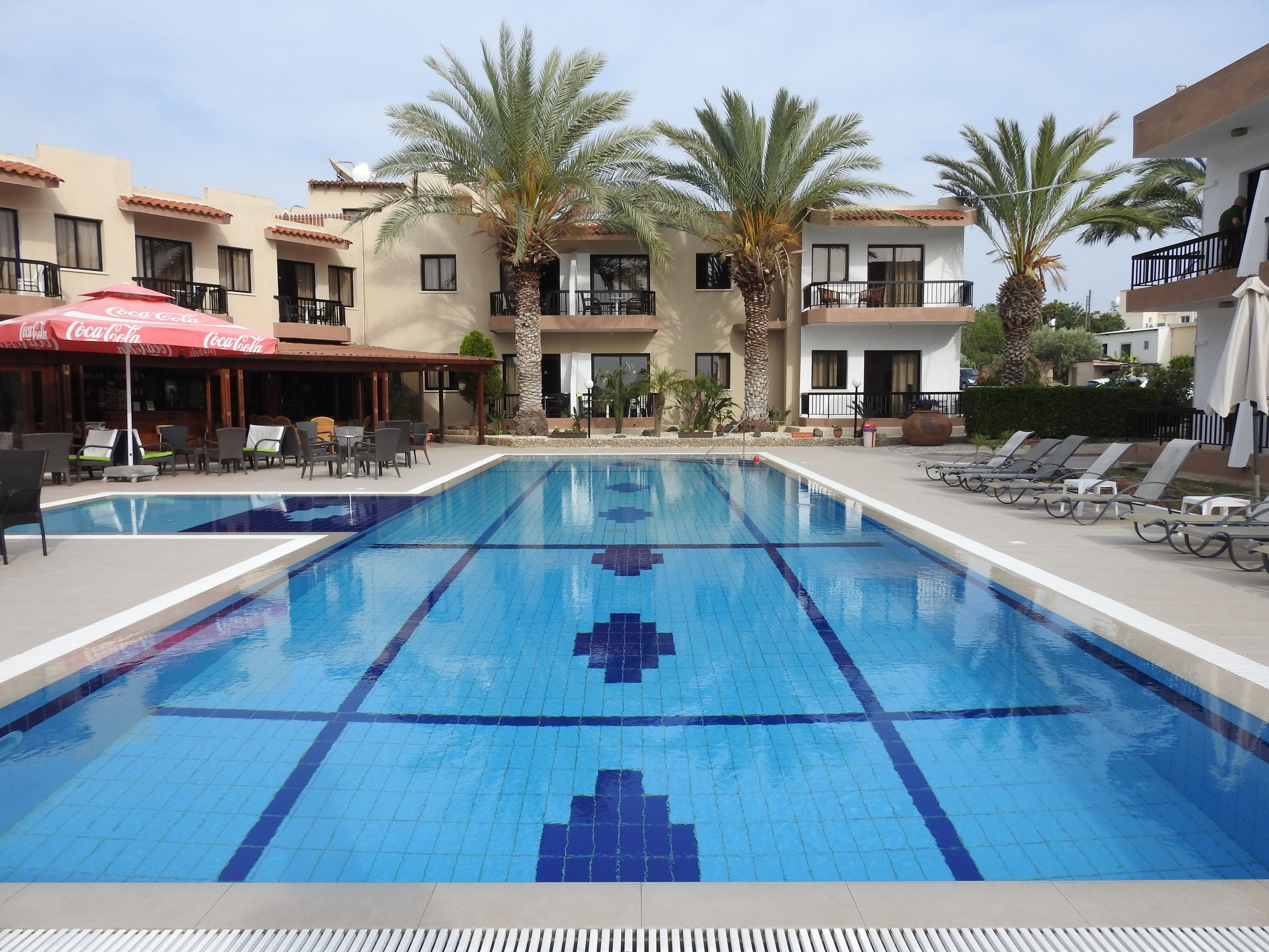 Une piscine dont le fond est décoré de motifs en mosaïque, entourée de chaises longues et de parasols, avec des palmiers et un bâtiment en arrière-plan.