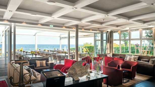 A spacious indoor area with a view of the sea, featuring red and beige seating, a large anchor decoration, and a wooden table. - Strandhotel Ostseeblick (Heringsdorf)