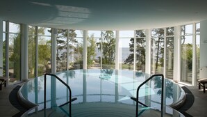 An indoor swimming pool with clear glass walls, surrounded by large windows offering views of trees and a body of water. - Strandhotel Ostseeblick (Heringsdorf)