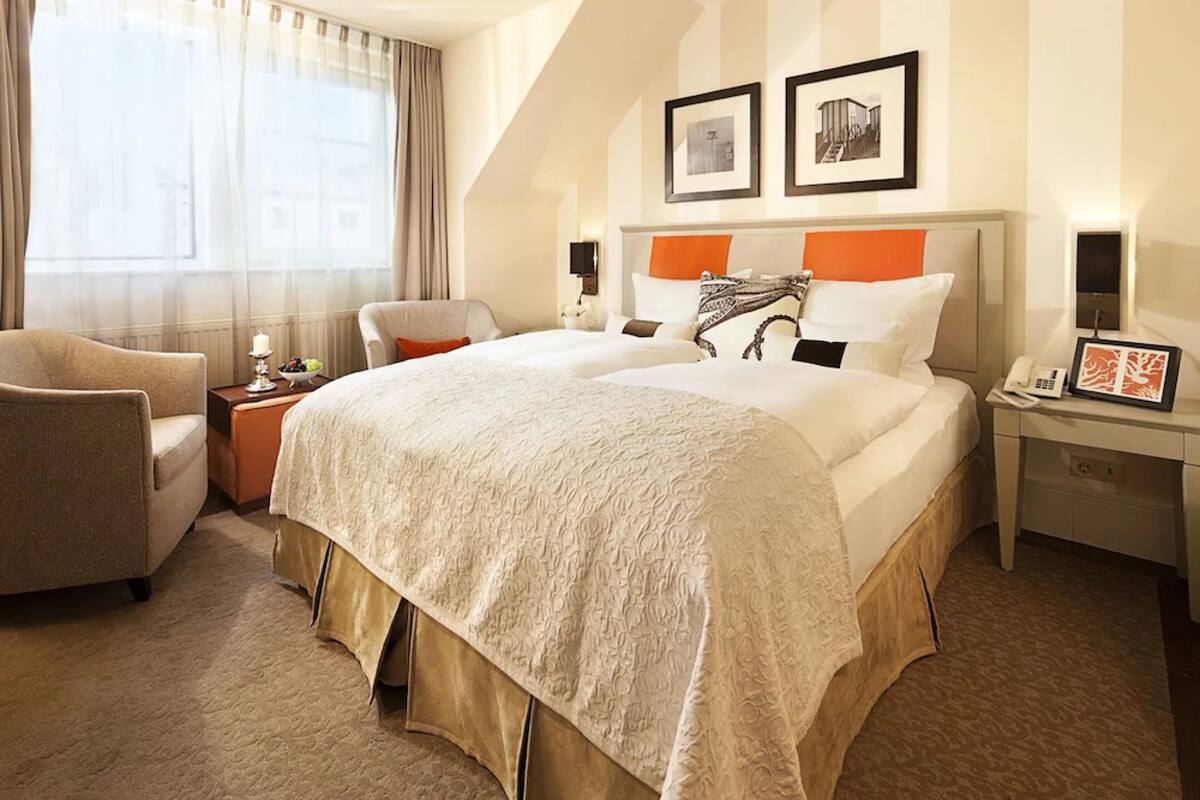 Habitación de hotel con una cama grande, una silla, una mesita de noche con lámpara y una ventana con cortinas.
