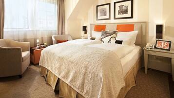 Quarto de hotel com uma cama grande, uma cadeira, uma mesinha de cabeceira com abajur e uma janela com cortinas.