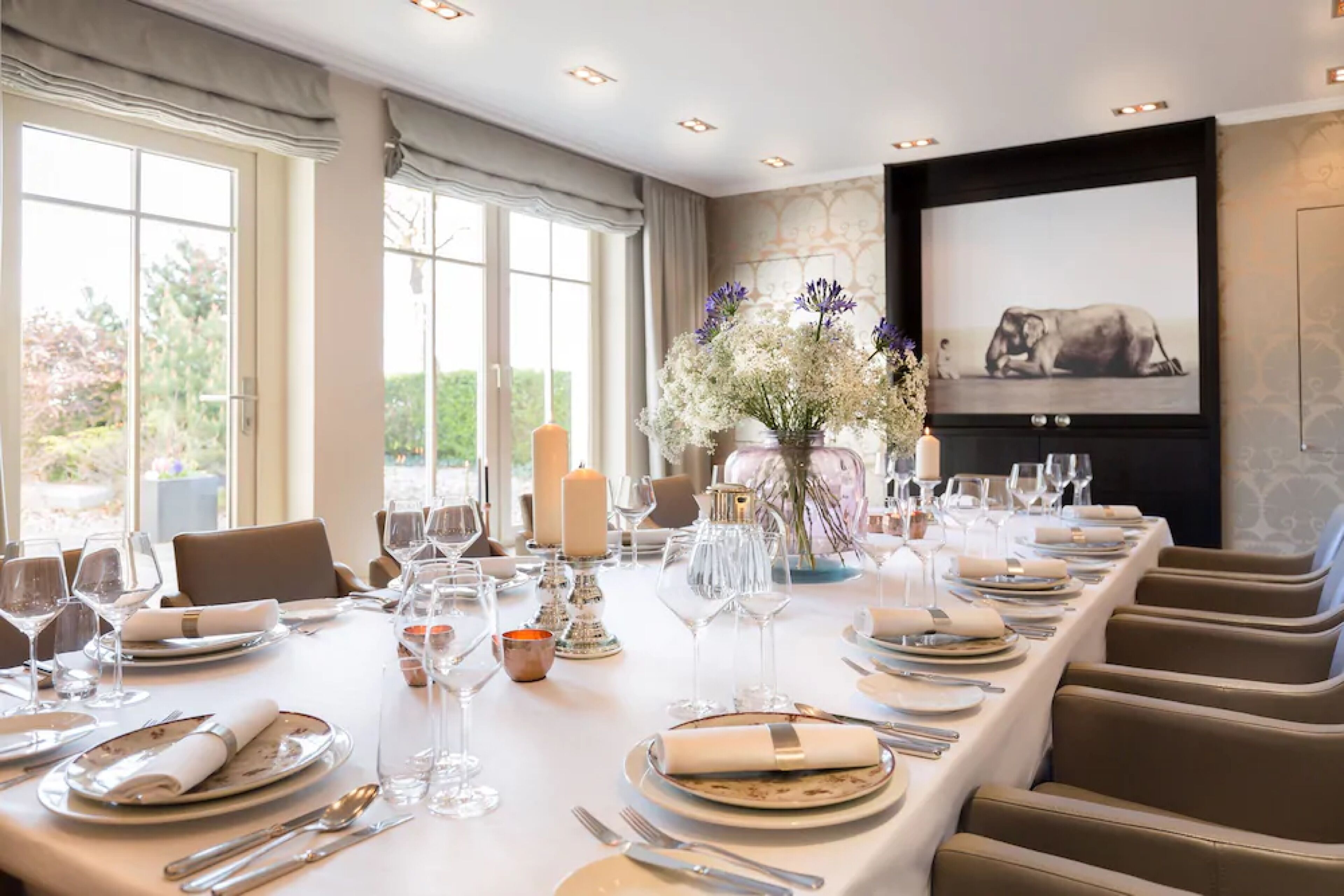 Une salle à manger élégante, avec une longue table dressée pour un repas, de grandes fenêtres et une œuvre d’art encadrée accrochée au mur.